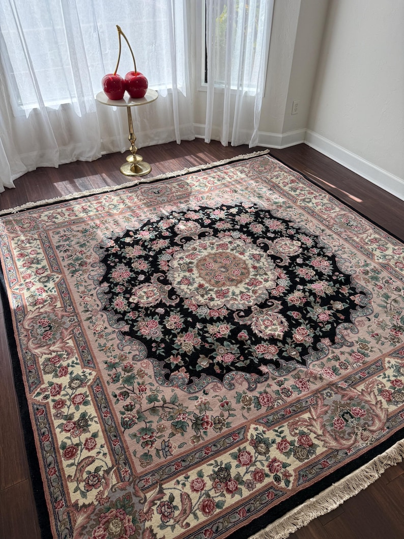 May include: A large, rectangular area rug with a central black medallion design featuring floral patterns in pink, cream, and green. The rug has a cream-colored border with floral motifs and a fringed edge. The rug is placed on a dark wood floor.
