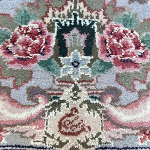 May include: Close-up of a detailed rug with a floral pattern. The rug features a central design with two large, red and pink peony-like flowers. The background is a mix of light blue, green, and beige tones, creating a soft, textured appearance.