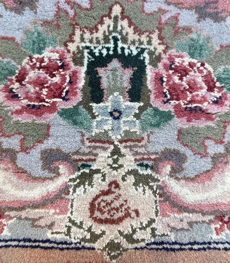 May include: Close-up of a detailed rug with a floral pattern. The rug features a central design with two large, red and pink peony-like flowers. The background is a mix of light blue, green, and beige tones, creating a soft, textured appearance.
