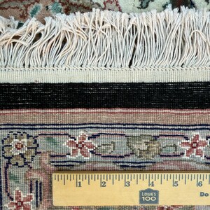 May include: Close-up of a section of an oriental rug with a floral pattern in shades of red, green, and beige. The rug has a cream-colored fringe and a black border. A wooden ruler is placed across the rug, showing measurements in inches.