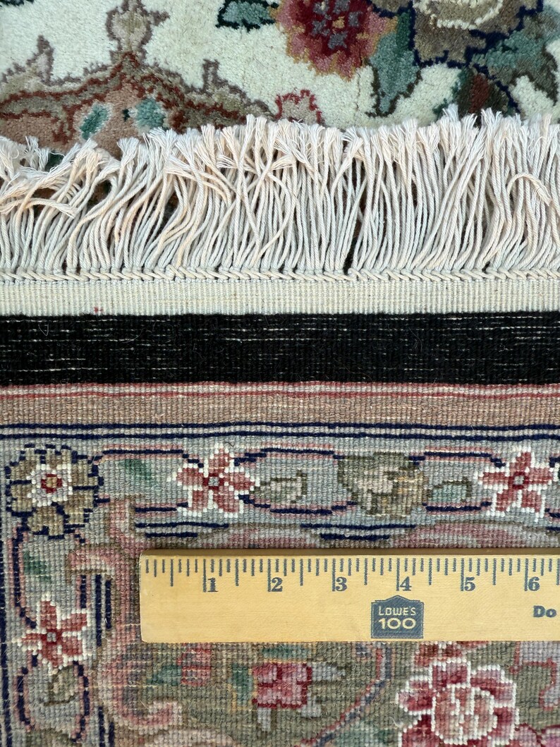 May include: Close-up of a section of an oriental rug with a floral pattern in shades of red, green, and beige. The rug has a cream-colored fringe and a black border. A wooden ruler is placed across the rug, showing measurements in inches.