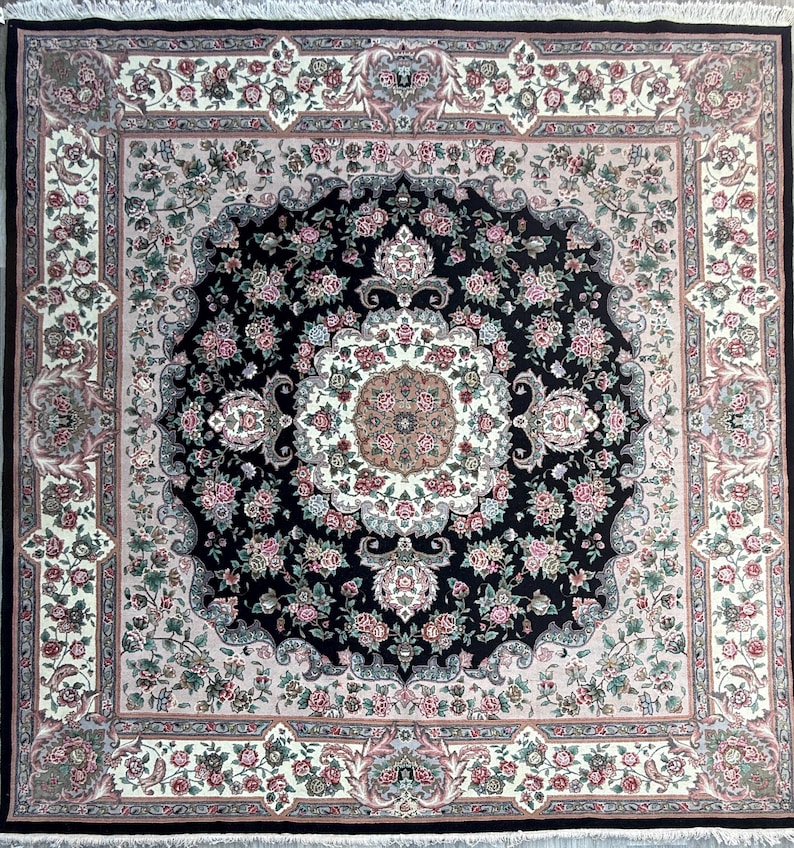 May include: Square area rug with a central black medallion design, surrounded by floral patterns in pink, cream, and green. The rug has a black border and fringe.