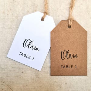 May include: Two tag-shaped place cards with twine strings. One is white, the other is brown. Both have the name "Olivia" in cursive and "TABLE 1" in a simple font. The tags are likely for a wedding or event.