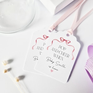 May include: Two white gift tags with pink ribbon designs. The text on the tags reads "Pop and Cheer When Baby Smith is here".