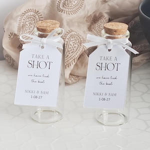 May include: Two clear glass bottles with cork stoppers and white tags. The tags read "TAKE A SHOT, we have tied the knot, NIKKI & SAM, 1-08-27". Each bottle is tied with a white ribbon. The background includes a patterned fabric.