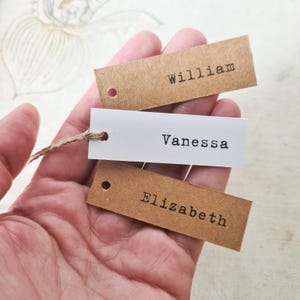 May include: Three rectangular gift tags with names printed on them. Two tags are brown, and one is white. The names printed are William, Vanessa, and Elizabeth. The tags are held together with twine.