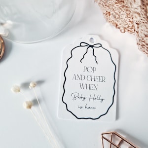 May include: White gift tag with black text that reads "Pop and cheer when Baby Holly is here". The tag has a black ribbon and a wavy border.