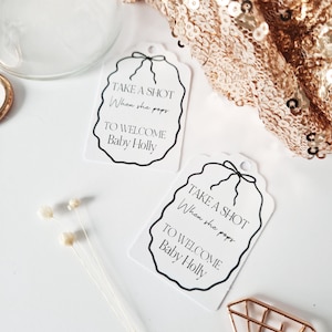 May include: Two white tags with black text and a black ribbon. The text on the tags reads "Take a shot when she pops to welcome Baby Holly".