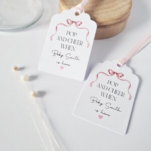 May include: Two white gift tags with pink ribbon designs. The text on the tags reads "Pop and Cheer When Baby Smith is here".