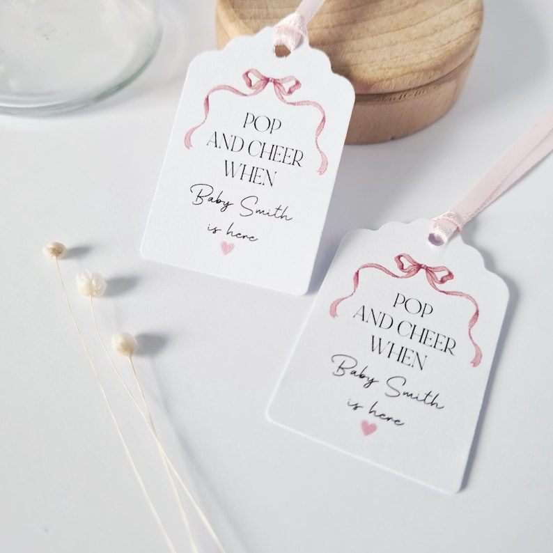 May include: Two white gift tags with pink ribbon designs. The text on the tags reads "Pop and Cheer When Baby Smith is here".