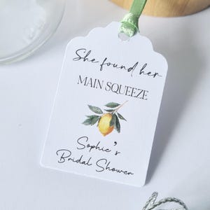 May include: White tag with scalloped edges and a green ribbon. The tag has black text that reads "She found her main squeeze, Sophie's Bridal Shower" and a watercolor illustration of a lemon on a branch.