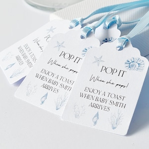May include: White gift tags with blue text and illustrations of seashells and starfish. The tags say "POP IT When she pops! Enjoy a toast when Baby Smith arrives." Each tag is tied with a light blue ribbon.