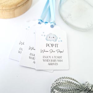 May include: White favour tags with blue ribbon and a blue cloud with a happy face and the text "POP IT When She Pops!" and "ENJOY A TOAST WHEN BABY SAM ARRIVES".