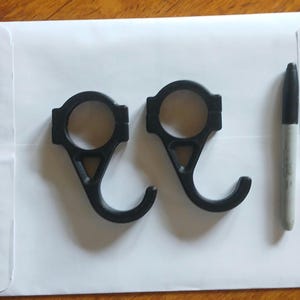 Puede incluir: Dos ganchos de plástico negro con una parte superior circular y una forma de gancho curvada. Un marcador negro está al lado de los ganchos. Los ganchos están sobre una superficie blanca.