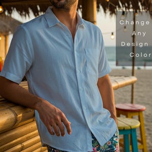 May include: A light blue short-sleeved button-down shirt with a pocket. The shirt is worn by a person leaning on a bamboo bar. The text on the image says "Change to Any Design or Color". The background includes a beach and cocktails.
