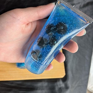 May include: A clear, flexible squeeze tube filled with a vibrant blue gel containing suspended dark, round, berry-like objects and fine glitter particles. The tube features a silver-toned crimped top seal and a matching blue screw-on cap at the base.