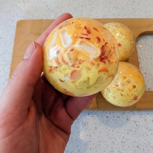 May include: A spherical, translucent bath bomb with a yellow and orange base, featuring embedded red and white decorative pieces throughout its surface. The object has a smooth, glossy finish and is held in a palm, with two additional identical spheres nearby.