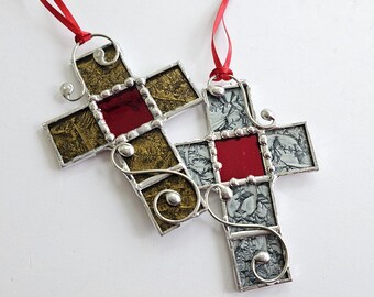 May include: Two stained glass cross ornaments with red and silver accents. One cross has a gold background and the other has a white background.