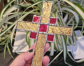 May include: A stained glass cross with gold textured glass and red accents. The cross is framed in silver and has a chain for hanging. The background features green and white striped leaves.