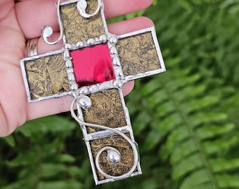 May include: A handcrafted stained glass cross ornament with a red center and gold-toned accents. The cross is trimmed with silver-colored metal and has decorative swirls. A red ribbon is attached for hanging.