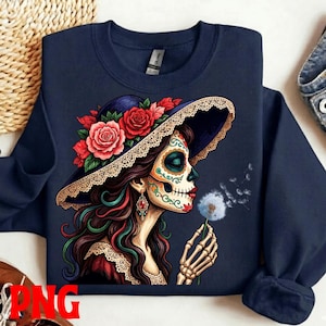 Könnte beinhalten: Marineblaues Sweatshirt mit einer farbenfrohen Illustration einer Frau mit Sugar-Skull-Make-up, die einen Hut mit roten Rosen trägt. Sie hält einen Löwenzahn, dessen Samen wegfliegen. Das Design ist detailliert und lebendig.