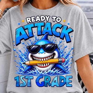 May include: A light grey t-shirt featuring a graphic of a blue shark wearing black sunglasses and holding a yellow pencil in its mouth. The text reads READY TO ATTACK 1ST GRADE, surrounded by blue water splashes and small yellow and blue stars.