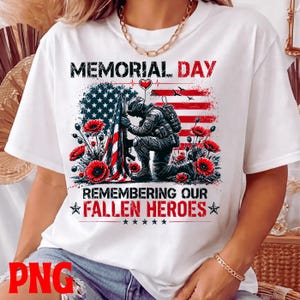 May include: White t-shirt with a Memorial Day design. The graphic shows a soldier kneeling before an American flag, surrounded by red poppies. Text reads "Memorial Day" and "Remembering Our Fallen Heroes."