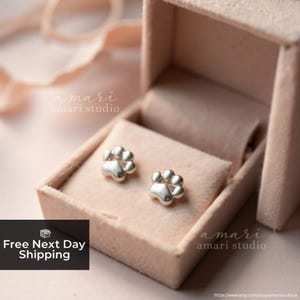 May include: A pair of silver paw print stud earrings displayed in a pink velvet jewelry box. The earrings are small and detailed, with a polished finish. The box is open, revealing the earrings on a soft, pink interior. The text "amari studio" is visible.