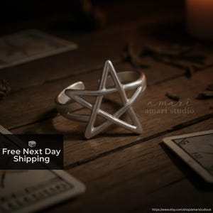 May include: A silver ring with a pentagram design. The ring is open at the back and sits on a dark wooden surface. The image includes the text "amari studio" and "Free Next Day Shipping".