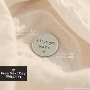 May include: A circular, pale grey pin featuring the words "I love you more" and a small heart. The pin is displayed on a soft, cream-coloured fabric. The words "amari studio" are printed on the fabric.