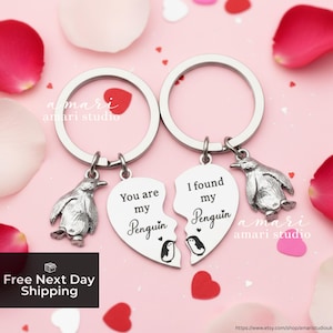 May include: Two silver keyrings with penguin charms and heart-shaped pendants that read "You are my Penguin" and "I found my Penguin". The background is pink with rose petals and small red hearts. The text "Free Next Day Shipping" is also visible.