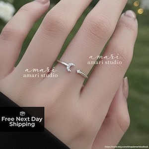 May include: A silver ring featuring a crescent moon and a star design. The ring is open, with the moon on one side and the star on the other. The ring is on a finger, with the brand name "amari amari studio" in the background.