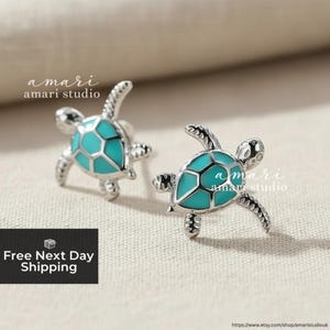 Könnte beinhalten: Silberne Schildkröten-Ohrstecker mit türkisfarbenen Emaille-Details. Die Ohrringe haben eine strukturierte Oberfläche und werden auf einem neutralen Hintergrund präsentiert. Der Markenname "amari studio" ist sichtbar.