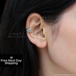 May include: A silver ear cuff with a geometric design is worn on the ear. The cuff has a series of parallel bars and a small roof-like structure. The text "amari amari studio" is visible on the image.