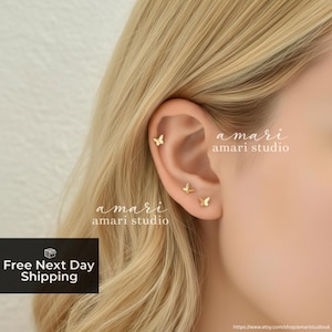 Puede incluir: Pendientes de mariposa dorados en una oreja. Los pendientes presentan un pequeño y delicado diseño de mariposa. La imagen también incluye el texto "amari amari studio".
