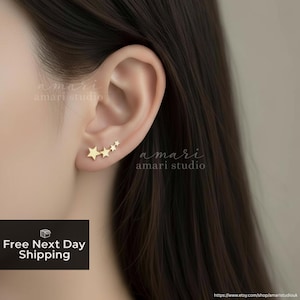 May include: Gold-coloured star-shaped earrings. The earrings are designed to climb up the ear, with four stars decreasing in size. The earrings are worn on a person's ear. The image also contains the text "amari studio".