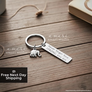 Op de afbeelding: Een zilveren sleutelhanger met een olifantenbedel en een rechthoekig label. Op het label staat "Never forget how STRONG you are". De sleutelhanger ligt op een houten oppervlak.
