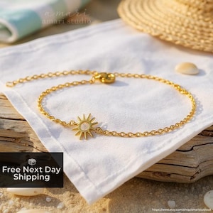 Puede incluir: Una delicada pulsera dorada con un dije de sol. La pulsera se muestra sobre una tela blanca, con un sombrero de paja y conchas marinas en el fondo. La cadena es fina y el dije de sol tiene un diseño detallado.