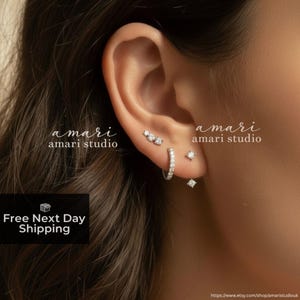 May include: A close-up of an ear adorned with multiple pieces of jewelry. The ear features a small hoop earring, a stud earring, and a diamond-shaped stud. The text "amari studio" is visible in the image.