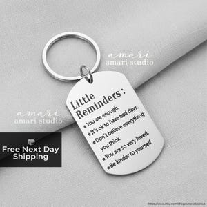 Peut inclure: Un porte-clés argenté avec un pendentif rectangulaire. Le pendentif porte les mots "Little Reminders" et des phrases inspirantes. Le porte-clés comprend un anneau argenté pour attacher les clés. Les mots "amari studio" sont également visibles.