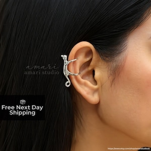 May include: A silver-colored ear cuff in the shape of a stylized cat, worn on the ear. The cuff wraps around the ear's cartilage, with the cat's body and tail forming the design. The image includes the text "amari studio".