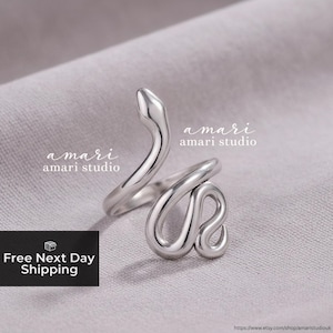 Puede incluir: Anillo plateado con forma de serpiente, con cuerpo enrollado y cabeza levantada. El anillo está sobre una tela gris. Se ve el texto "amari amari studio".