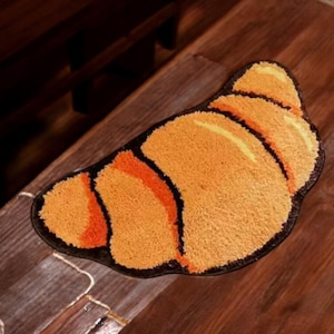 Puede incluir: Una alfombra decorativa con forma de croissant. La alfombra presenta un diseño texturizado en naranja y amarillo con detalles en naranja oscuro y un borde negro. La alfombra está colocada sobre una superficie de madera.