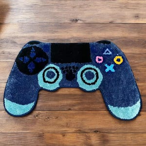 Könnte beinhalten: Ein blauer Teppich in Form eines Videospiel-Controllers. Der Teppich hat ein schwarzes Mittelteil, ein blaues Steuerkreuz und bunte Knöpfe. Das Design des Controllers umfasst zwei hellblaue Kreise und eine hellblaue Basis.