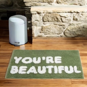 Op de afbeelding: Een groene rechthoekige badmat met de woorden "YOU'RE BEAUTIFUL" in het wit, op een houten vloer. Links van de mat staat een witte vuilnisbak. De achtergrond toont een stenen muur en een houten richel.