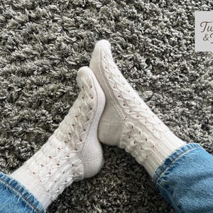 Könnte beinhalten: Ein Paar weiße Zopfstricksocken mit Lochmuster, getragen mit blauen Jeans. Die Socken werden auf einer grauen, strukturierten Oberfläche gezeigt. Das Logo "Twist & Toe" befindet sich oben rechts.
