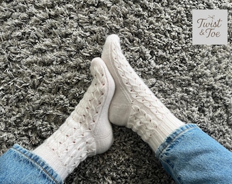 May include: Cream-coloured cable knit socks with openwork patterns, worn with blue denim jeans. The socks are shown on a grey, textured surface. The logo "Twist & Toe" is in the upper right corner.