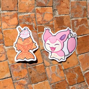 May include: Two stickers on a brick surface. One sticker is a cartoon fish-shaped pastry topped with ice cream and a straw. The other sticker is a pink cartoon character with a smiling face and a long tail.