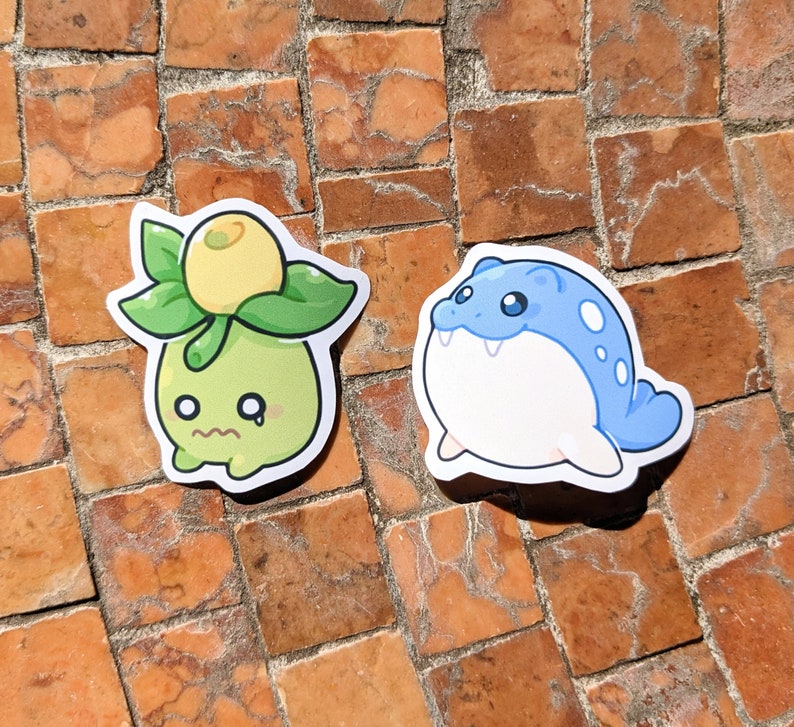 May include: Two die-cut stickers featuring cartoon characters. One is a green plant-like creature with a yellow fruit and sad expression. The other is a blue and white seal-like creature with a smiling face.