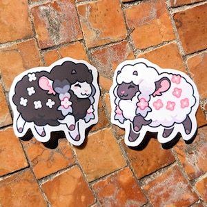 May include: Two sheep stickers, one black and one white, each adorned with floral accents. The black sheep features white flowers, while the white sheep has pink flowers. Both have pink bows and are set against a brick background.
